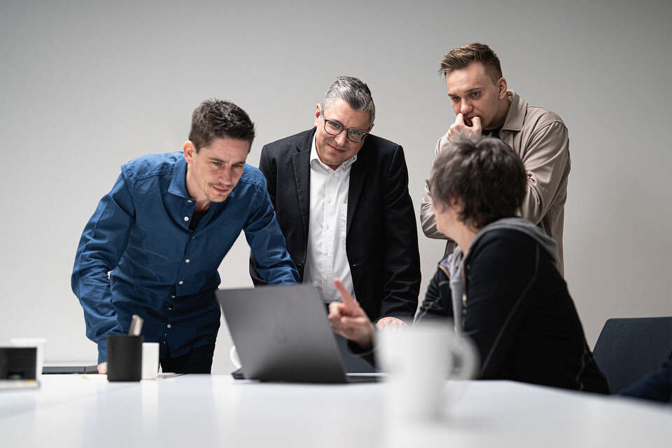 Drei Männer stehen um einen Tisch, eine Frau sitzt davor und zeigt auf einen Laptop auf dem Tisch
